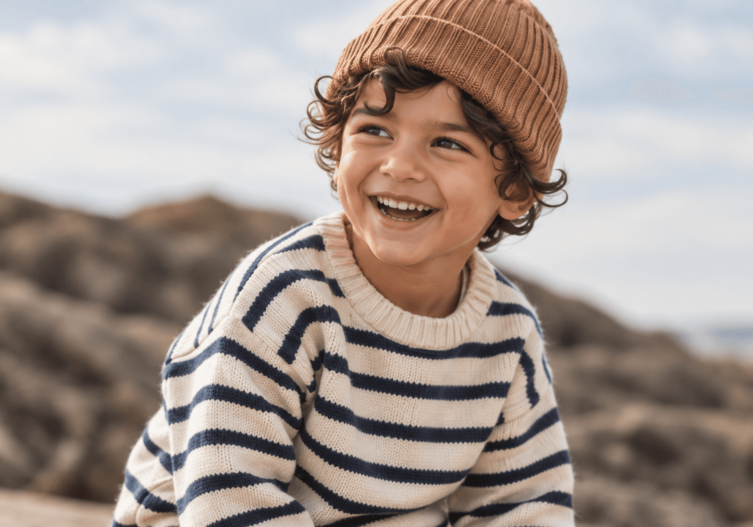 A smiling child by the coast in a striped sweater and a warm knitted hat